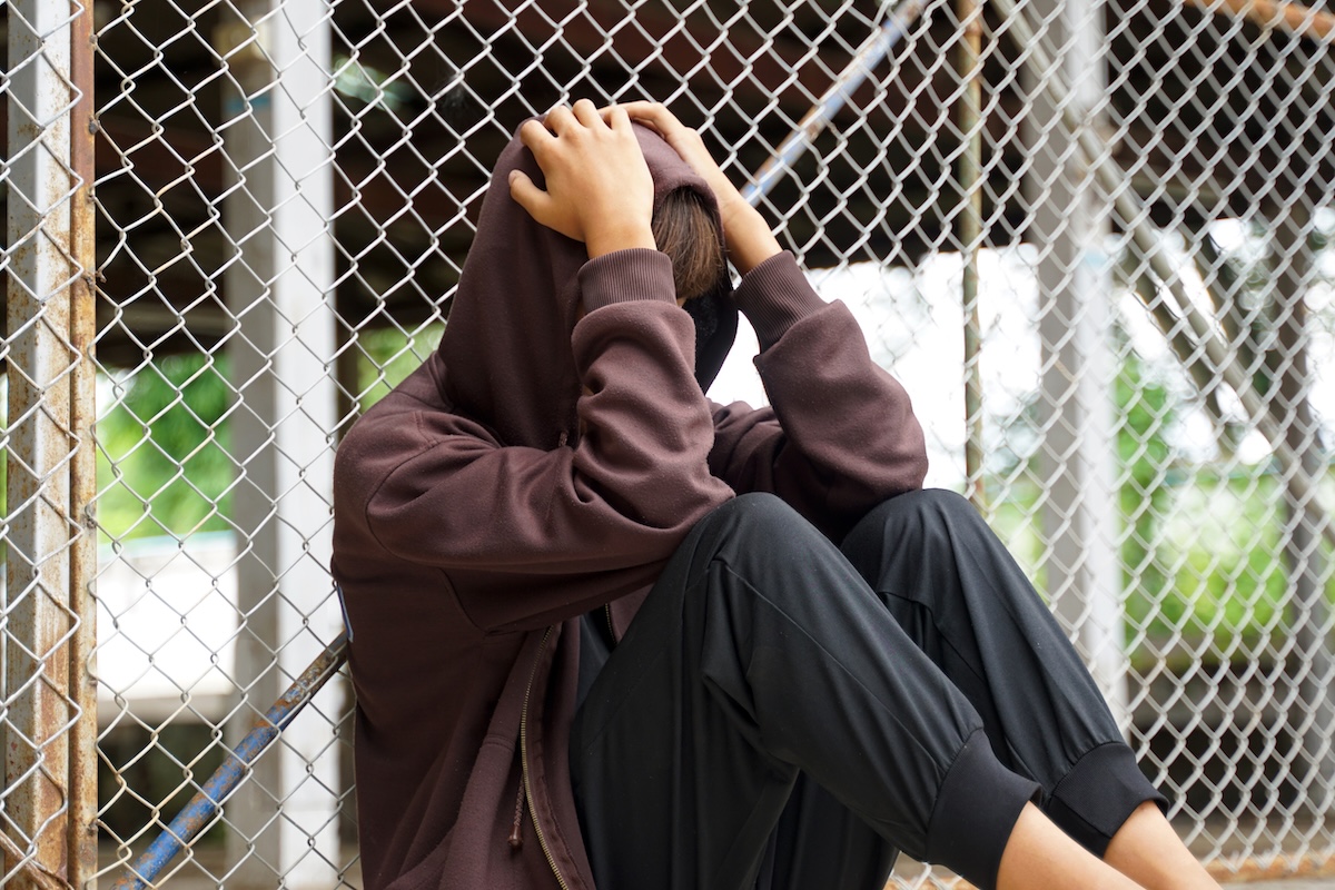 A young male wearing a hoodie sits with his face in his hands resting against a wire mesh fence