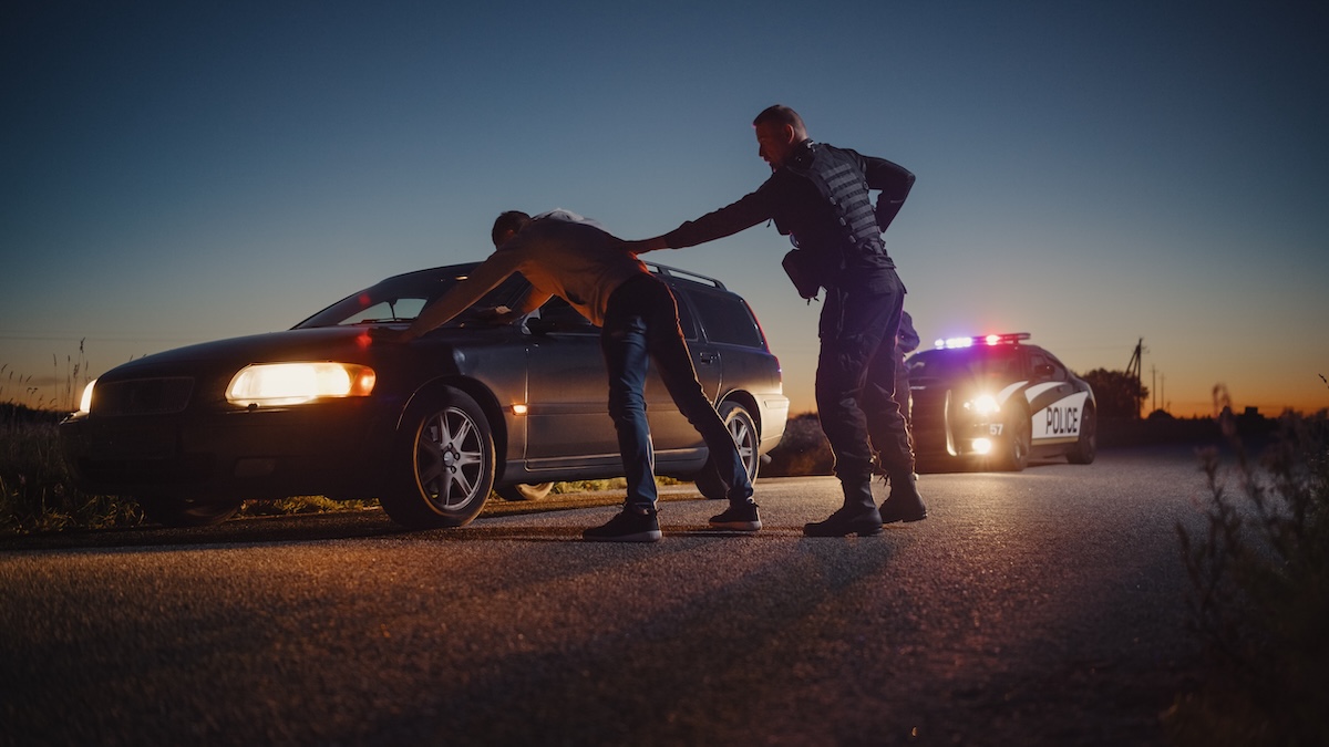 A Policeman Performing a Pat-Down Search on a Man With his Hands on Car Hood.