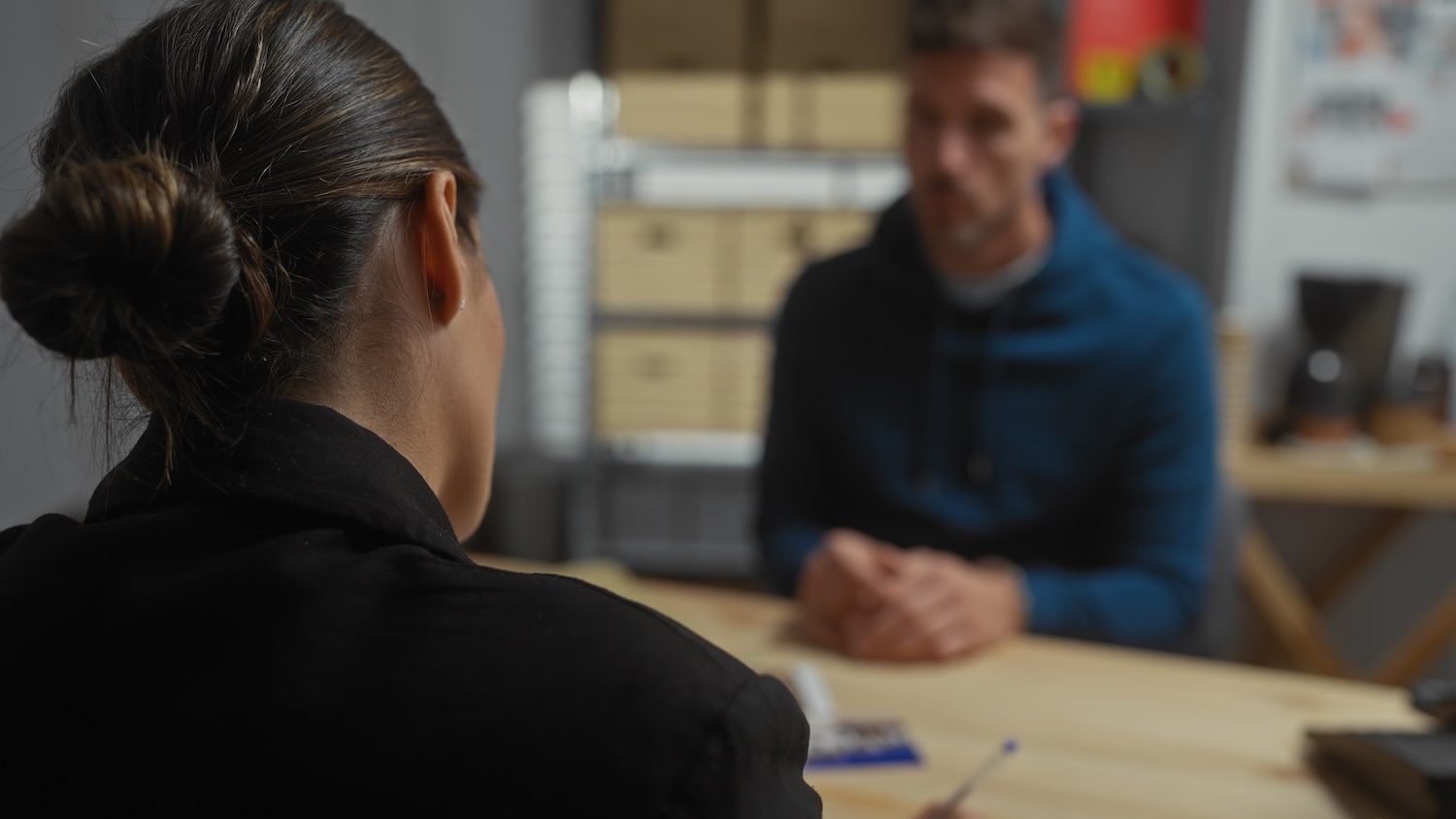 A woman with dark hair in a bun sits at a desk facing a man in a blue hoodie, having a conversation in an office setting. The focus is on the woman’s back, with the man blurred in the background.