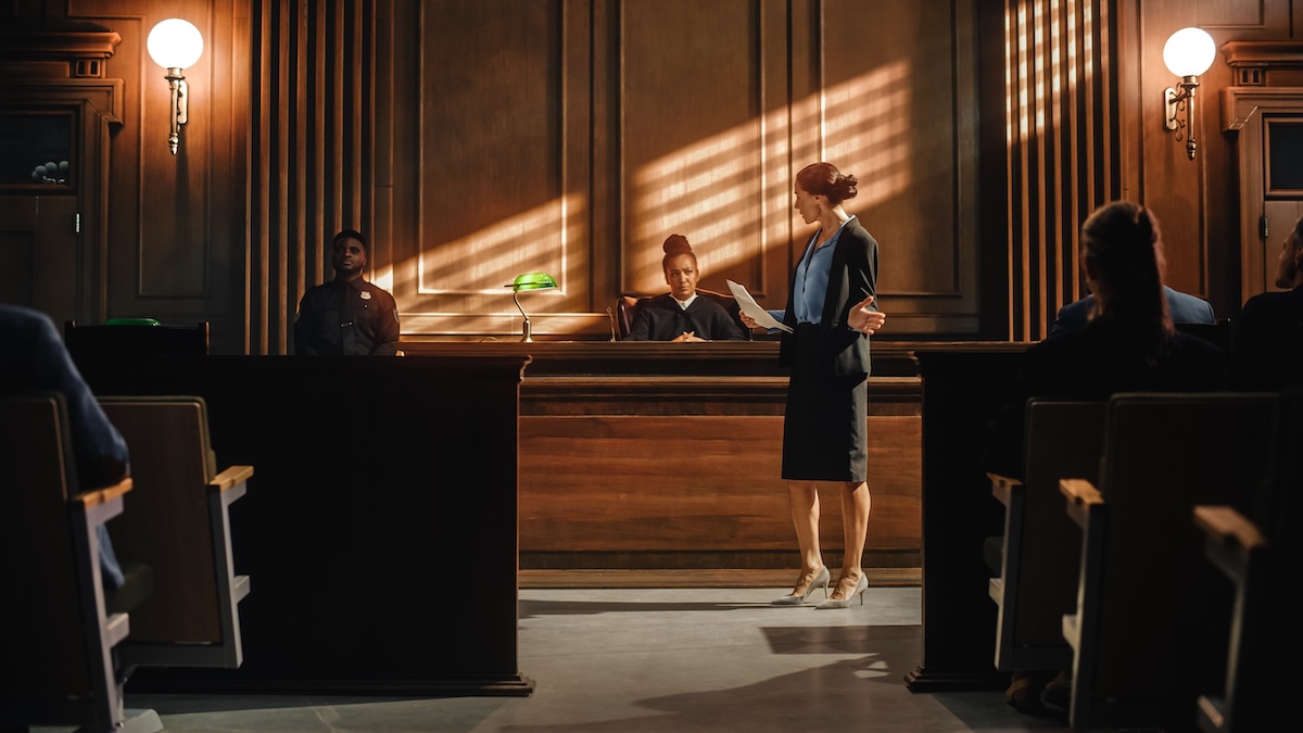 A lawyer stands and speaks in a courtroom before a judge seated at the bench, with a court officer nearby. Sunlight streams through windows, casting lines of light across the wooden interior.