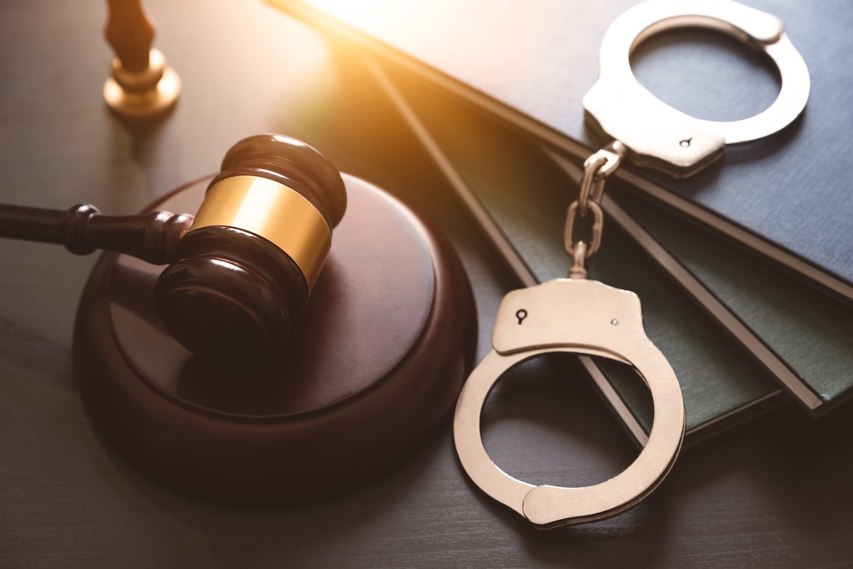 A judges gavel rests on a wooden table next to an open law book and a pair of metal handcuffs, symbolizing law, justice, and criminal proceedings. Sunlight shines on the items.