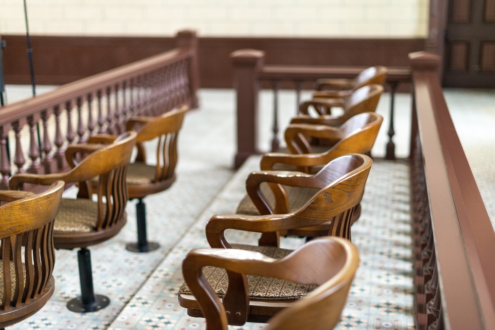 A row of empty wooden jury chairs with armrests in a courtroom, lined up behind a wooden railing on a patterned tile floor.