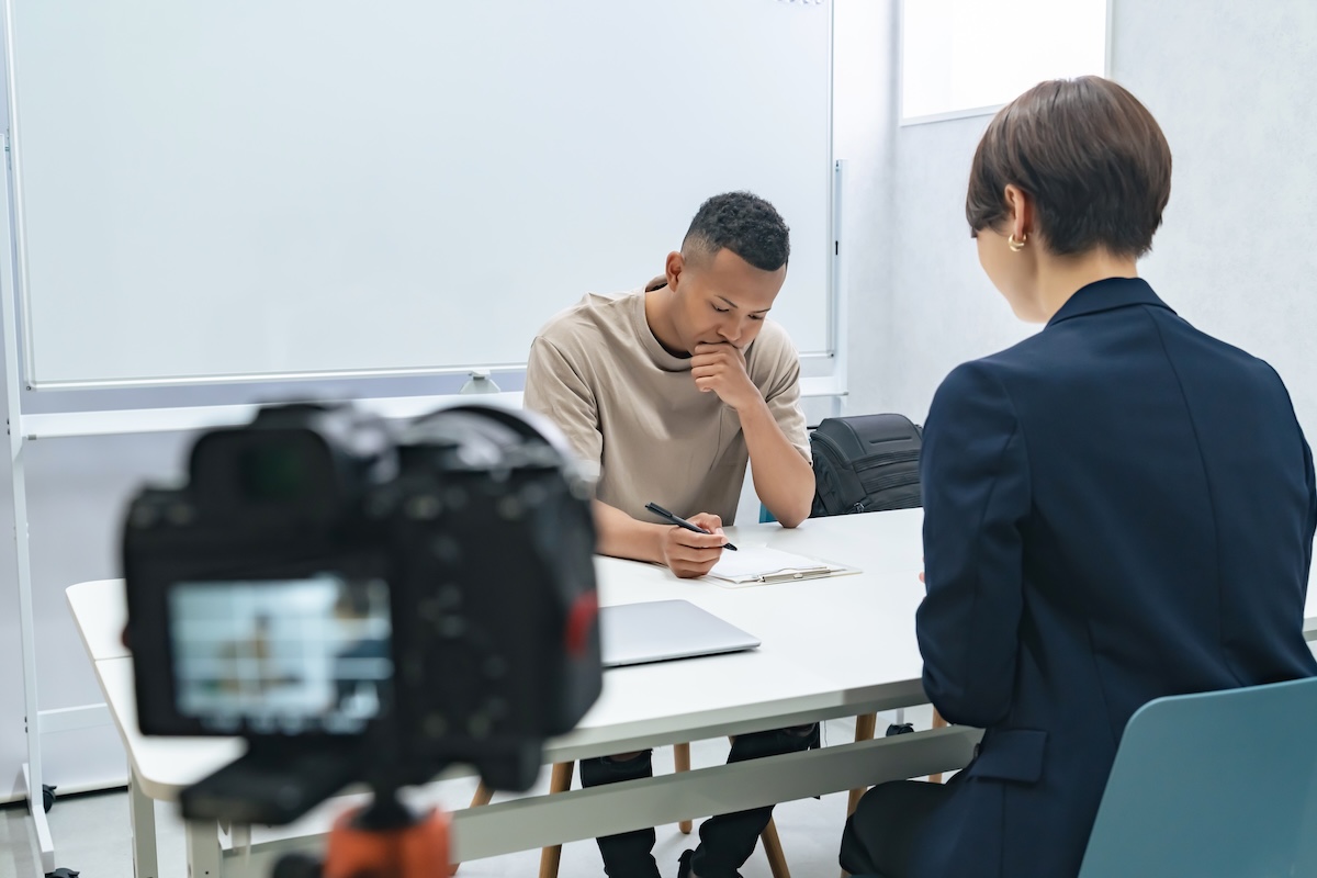 A young man sits at a table across from a woman in an office setting, both focused on paperwork, with a camera set up in the foreground recording the scene.