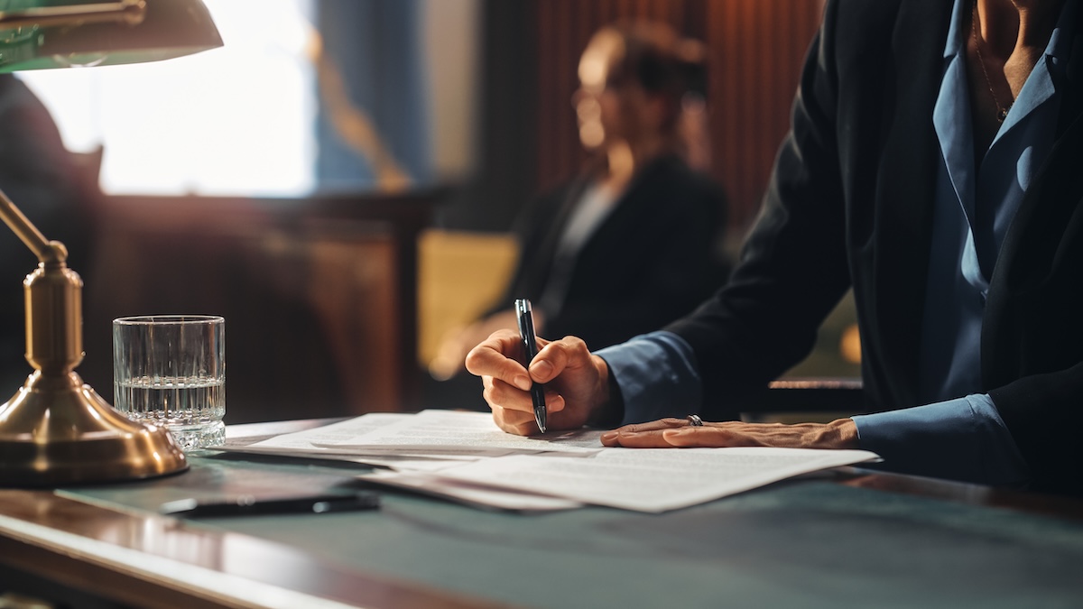 A person in a suit sits at a desk, writing on documents with a pen. A glass of water, a lamp, and more papers are on the desk. Another person sits blurred in the background, suggesting a formal setting like a courtroom or office.