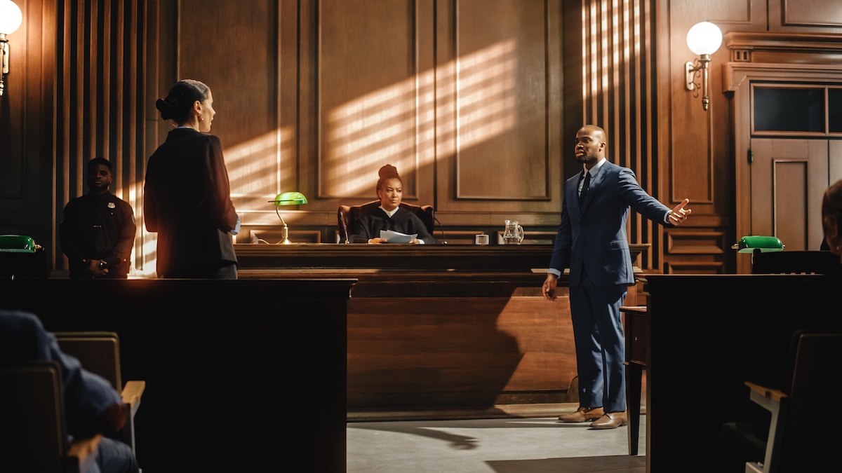 A lawyer in a blue suit speaks passionately in a courtroom, facing a woman while a judge listens from the bench. Another person stands near the wall, and warm light filters into the wood-paneled room.
