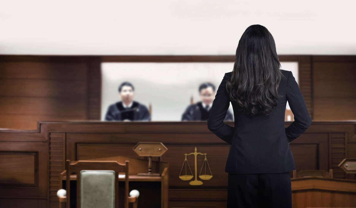 A woman in a black suit stands in a courtroom, facing two judges seated behind a bench. The setting is formal, with wooden furniture and a symbol of justice visible.