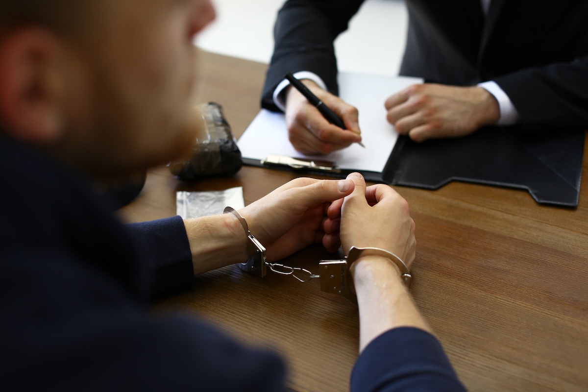 A person with handcuffed wrists sits at a table across from another individual in a suit who is writing on a clipboard. Various objects, including a pen, are on the table.