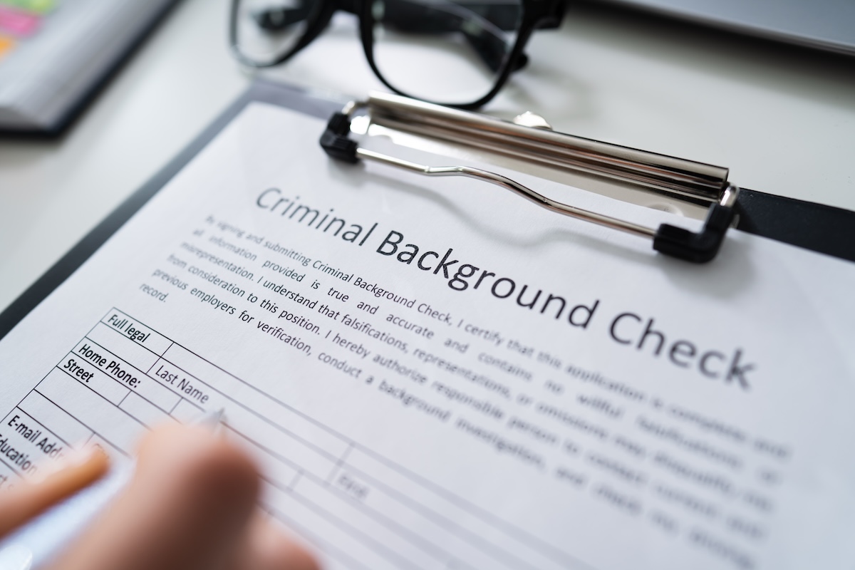 A close-up of a clipboard holding a Criminal Background Check form, with a hand filling it out. Glasses are placed nearby on a white surface.
