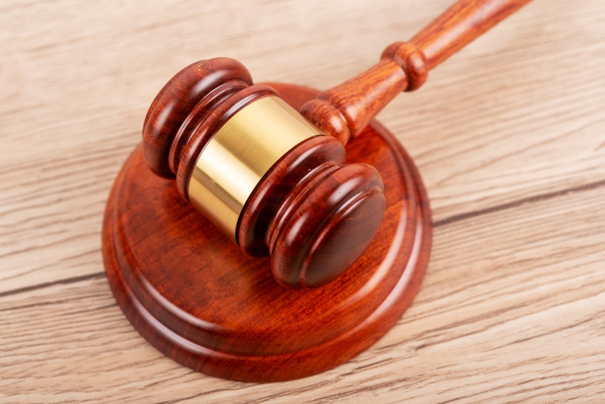 A close-up of a wooden judges gavel resting on a matching sound block, placed on a light wooden surface. The gavel has a brass band around the head.