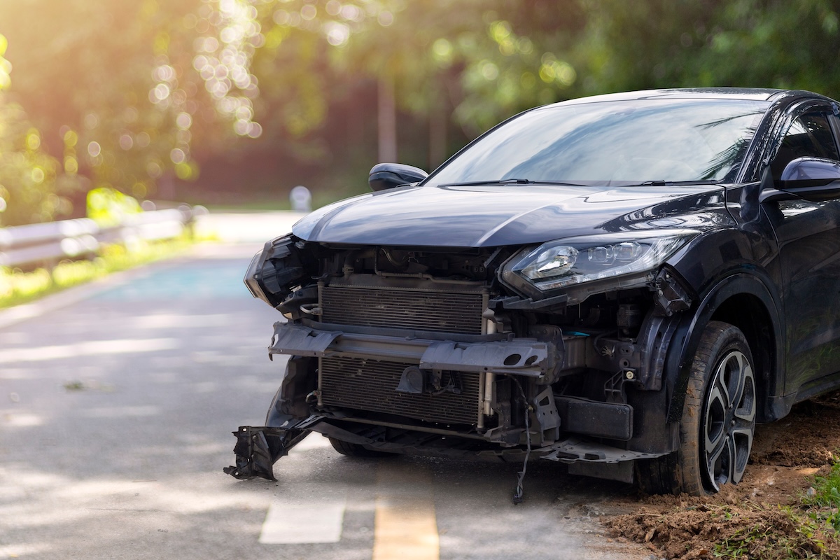 A black car with severe front-end damage is parked on the side of a road. The front bumper and grille are missing, exposing the internal components. Trees and greenery are visible in the background.