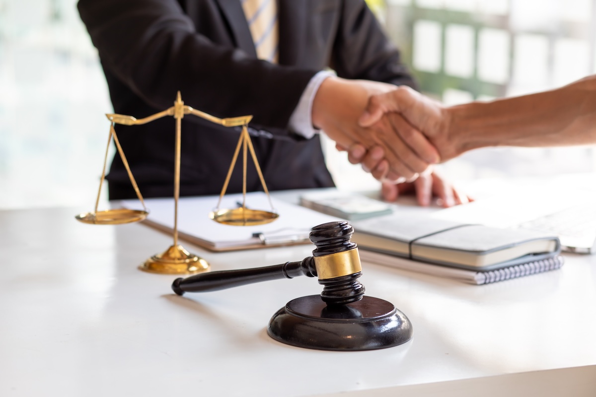 A judge’s gavel and scales of justice sit on a desk while two people in business attire shake hands in the background, suggesting a legal agreement or conclusion to a case.