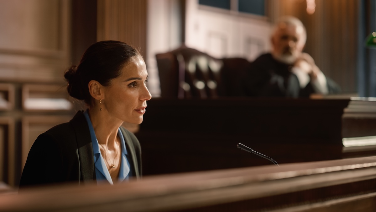 A woman in professional attire speaks into a microphone while seated in a courtroom witness stand, with a judge observing from the bench in the background.