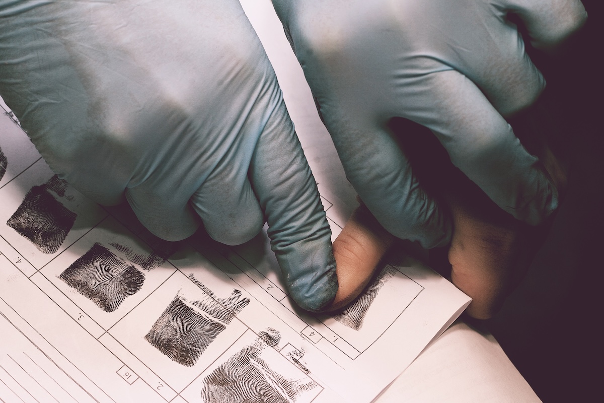 A person wearing blue gloves presses another persons finger onto an ink sheet to take fingerprints, with fingerprint samples visible on the paper.