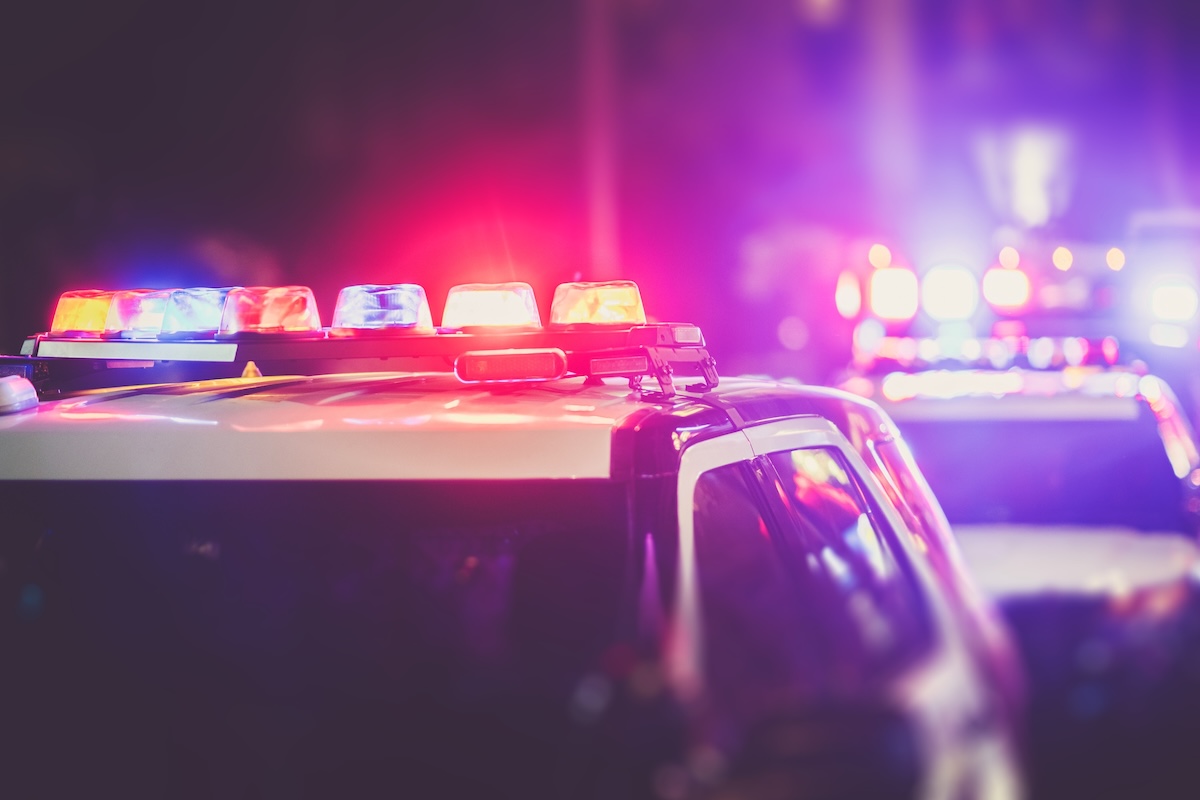 Police cars with flashing red and blue lights are parked on a street at night, creating a dramatic, blurred scene with emergency lights illuminating the area.