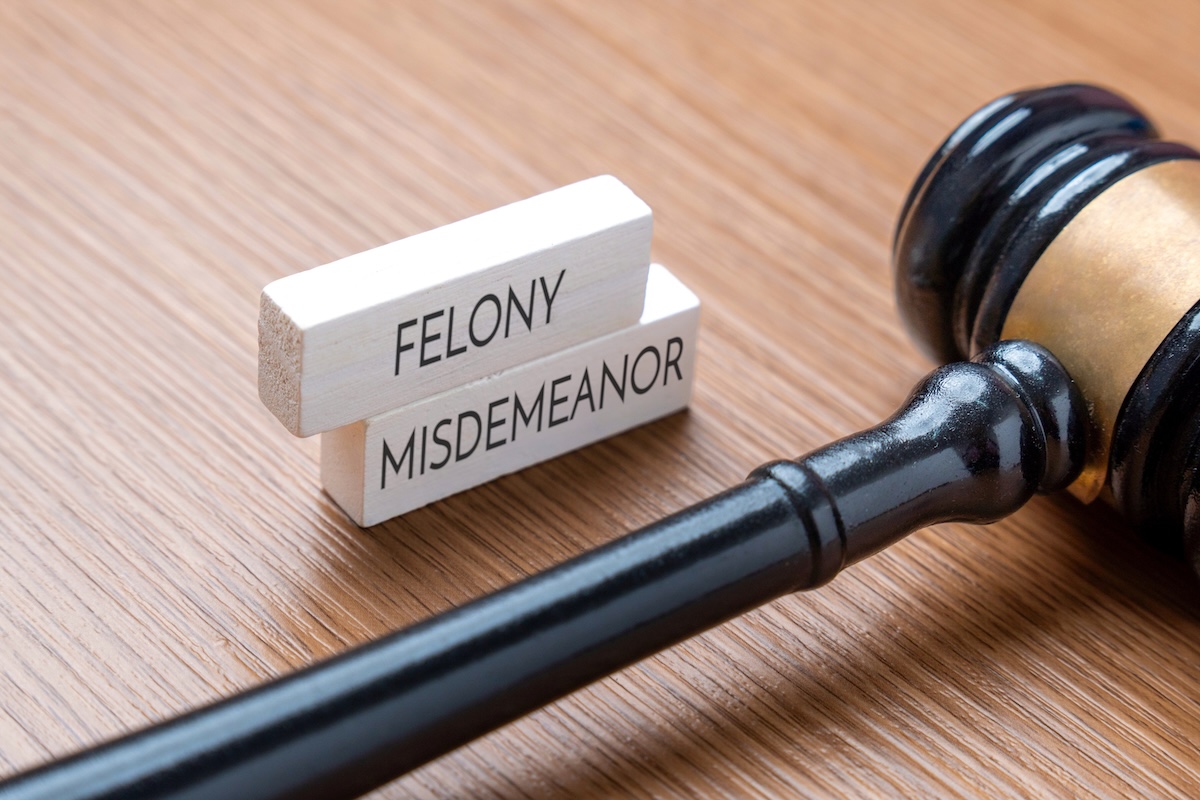 A judge’s gavel lies on a wooden surface next to two stacked wooden blocks labeled FELONY and MISDEMEANOR, representing different levels of criminal offenses.