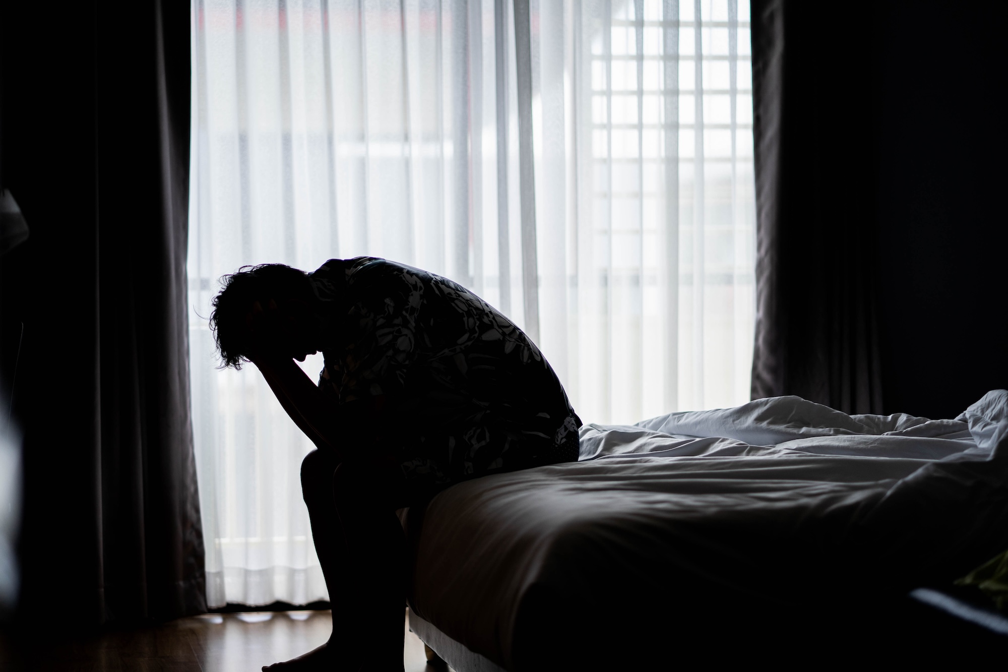 A person sits on the edge of a bed in a dimly lit room, head in hands, silhouetted against a window with sheer curtains, suggesting feelings of sadness or distress.
