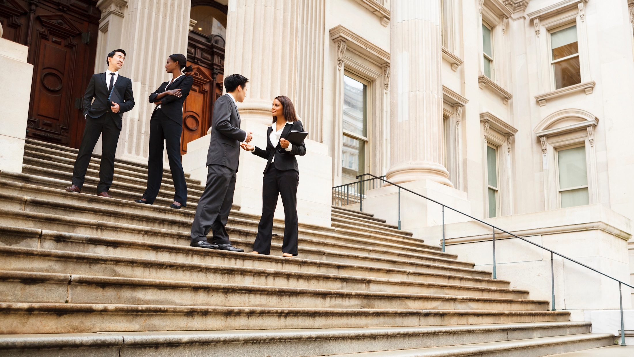 Four people in business attire stand and converse on the steps of a large, classical-style building with columns, possibly a courthouse or government building.