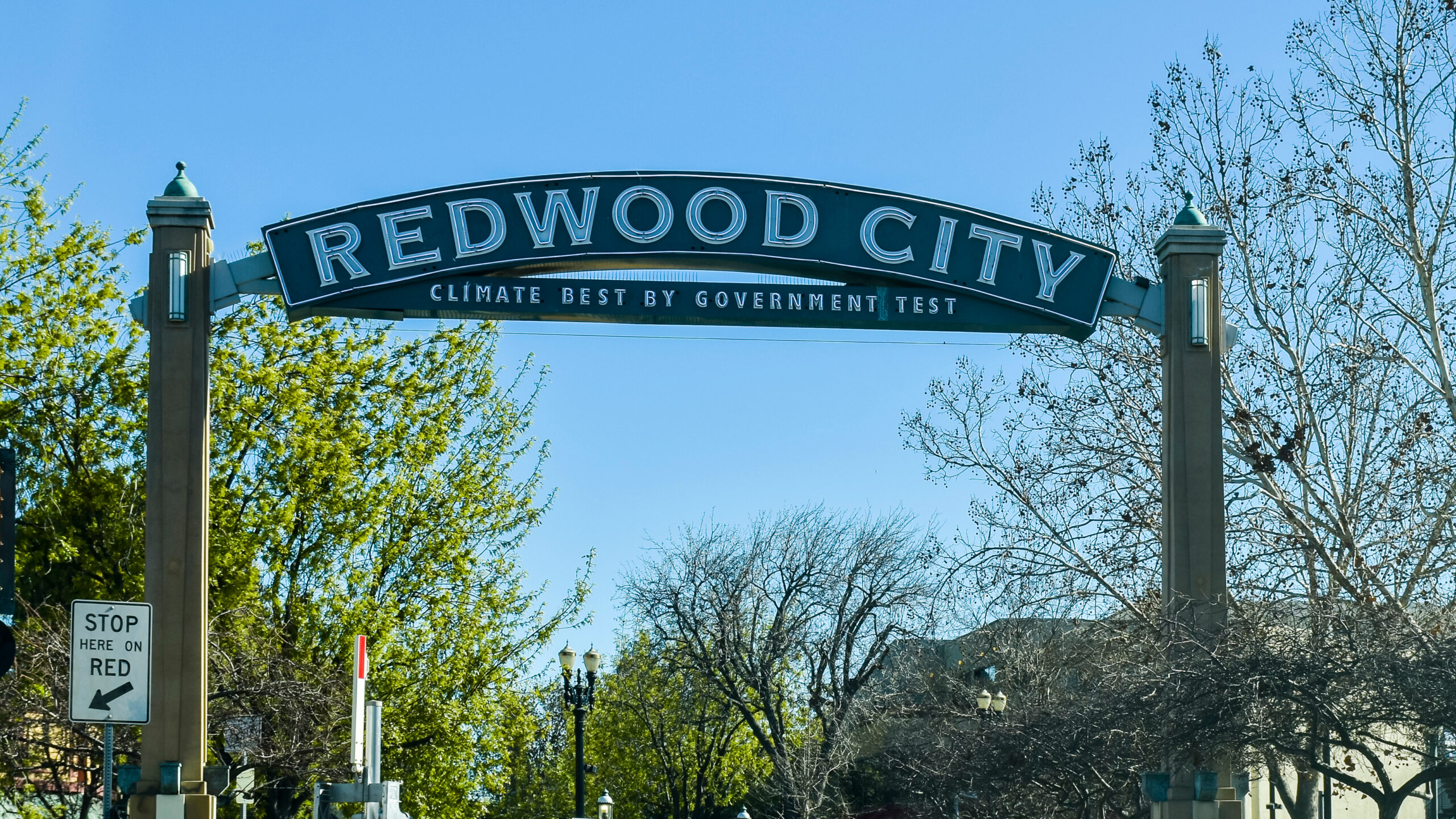 A large sign reads REDWOOD CITY with the slogan CLIMATE BEST BY GOVERNMENT TEST below it. Trees with green and bare branches are visible under a clear blue sky. A STOP HERE ON RED sign is also present.