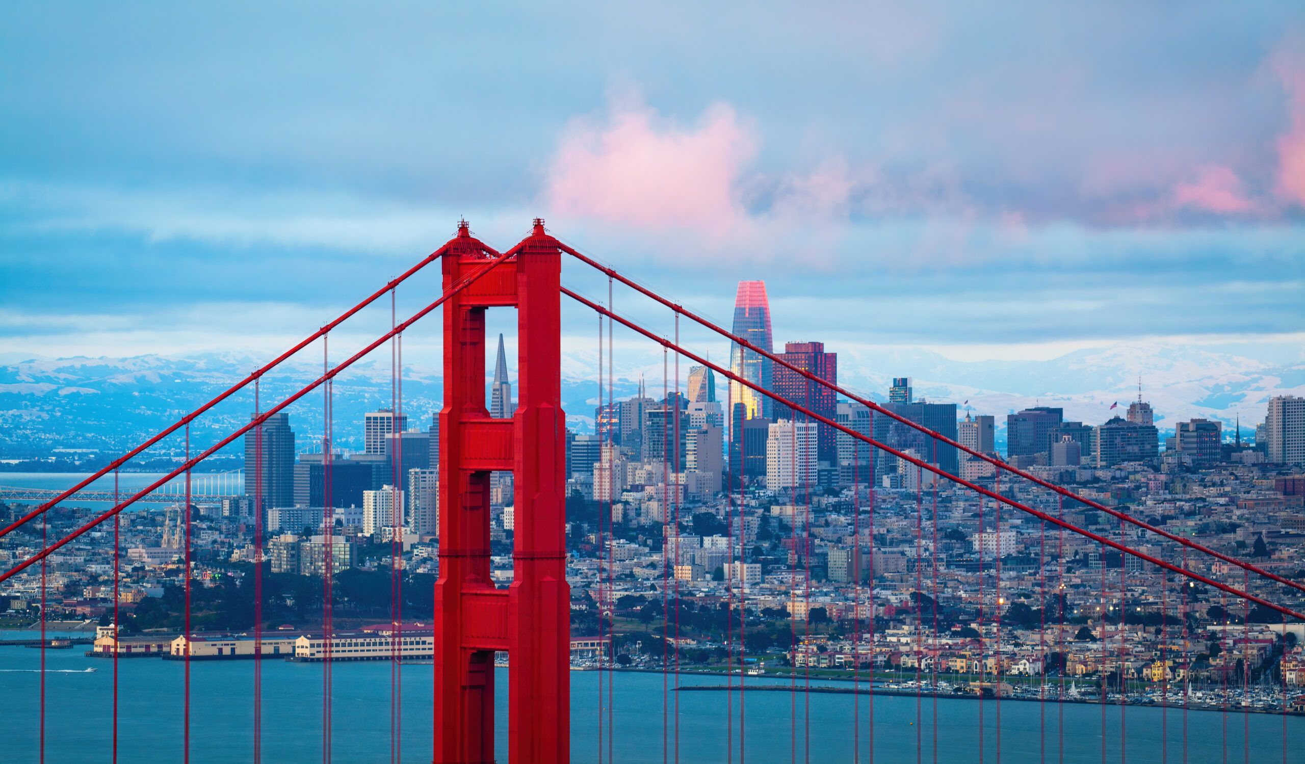 The Golden Gate Bridge’s red towers and cables frame a view of downtown San Francisco, with skyscrapers and buildings under a cloudy sky tinted with pink at sunset.