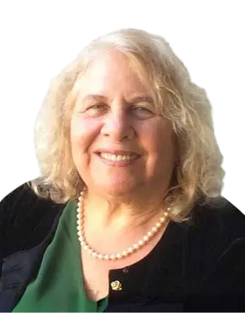 Smiling older woman with wavy gray-blonde hair, wearing a green top, black jacket, a pearl necklace, and a pin, against a plain light background.