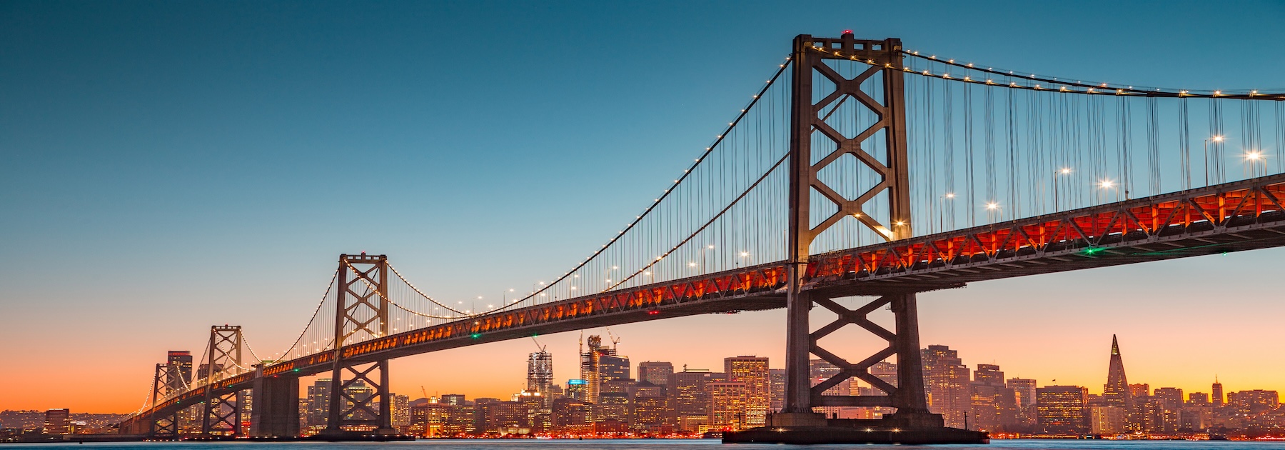 The Bay Bridge stretches across the water at sunset, with the San Francisco skyline illuminated in the background under a clear sky.