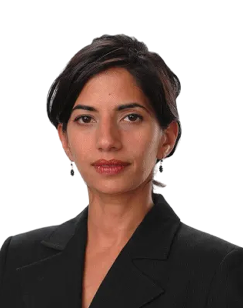 A woman with short dark hair wearing a black blazer and small drop earrings, posing against a plain white background.