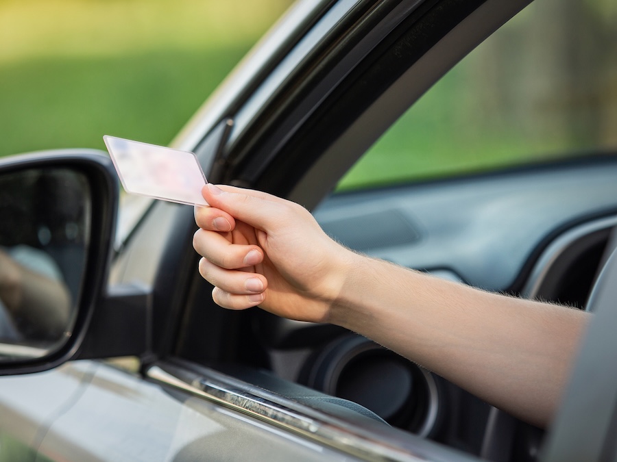 A person’s hand extends out of a car window holding a card, possibly a drivers license or ID, with a blurred background of greenery.