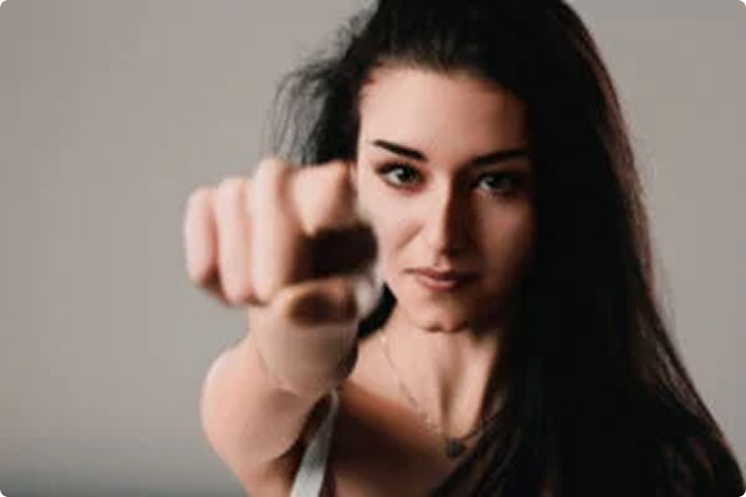 A woman with long dark hair faces the camera, extending her arm forward with a clenched fist, as if punching toward the viewer. She has a determined expression and wears a light-colored tank top. The background is plain and neutral.
