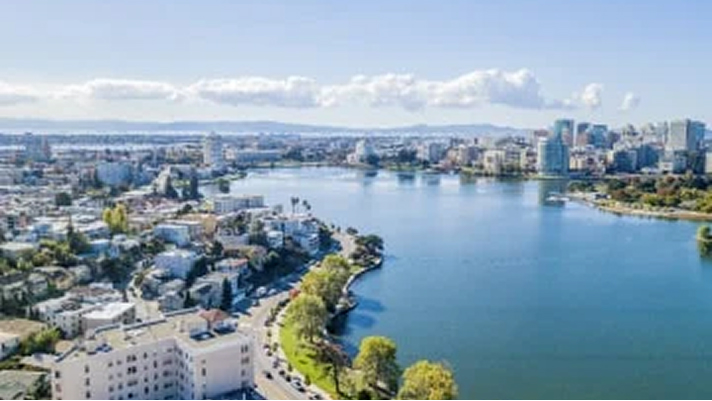Aerial view of a city with a large lake bordered by trees and buildings, under a clear blue sky with scattered clouds. High-rise and mid-rise buildings are visible in the distance along the waterfront.