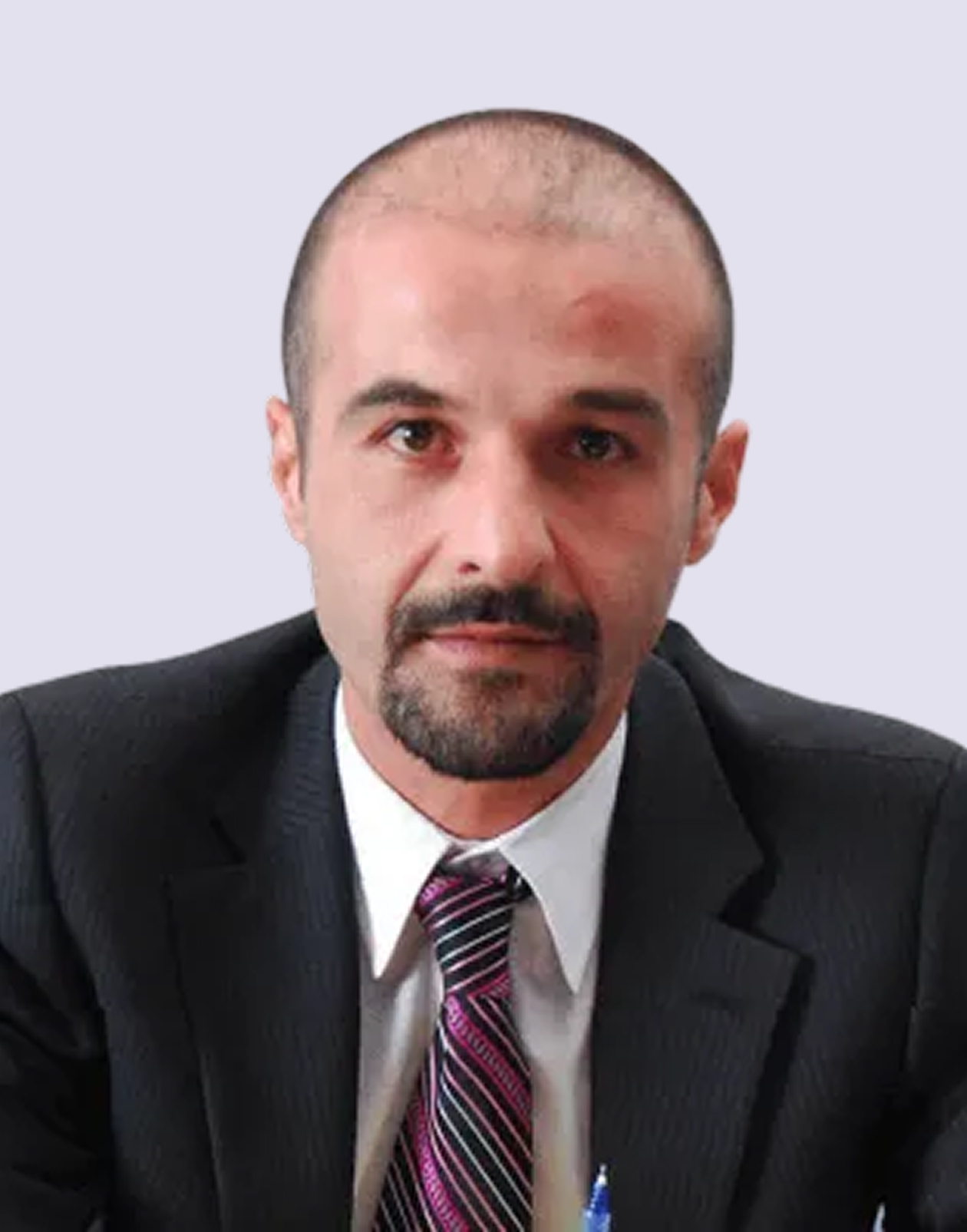 A man with a shaved head and goatee wearing a dark suit, white shirt, and striped tie, sitting against a plain light background.