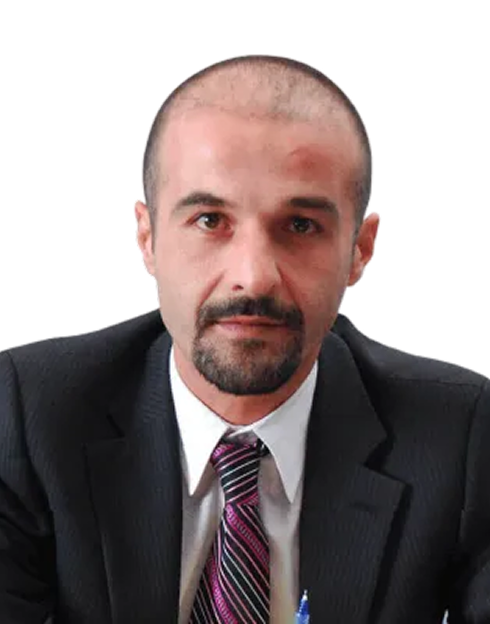 A man with a shaved head and goatee wearing a black suit, white shirt, and striped tie, looking directly at the camera against a plain light background.