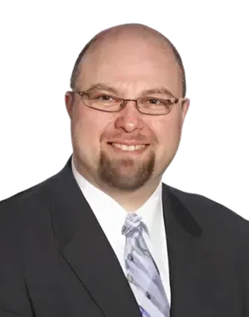 A man with glasses and a goatee is smiling, wearing a dark suit, white shirt, and a light-colored patterned tie against a plain light background.