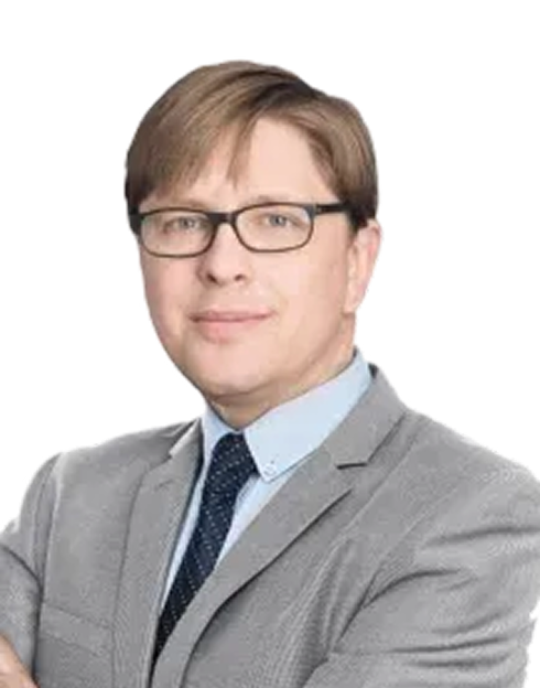 A man wearing glasses and a grey suit with a light blue shirt and dark tie, posing with his arms crossed against a plain white background.