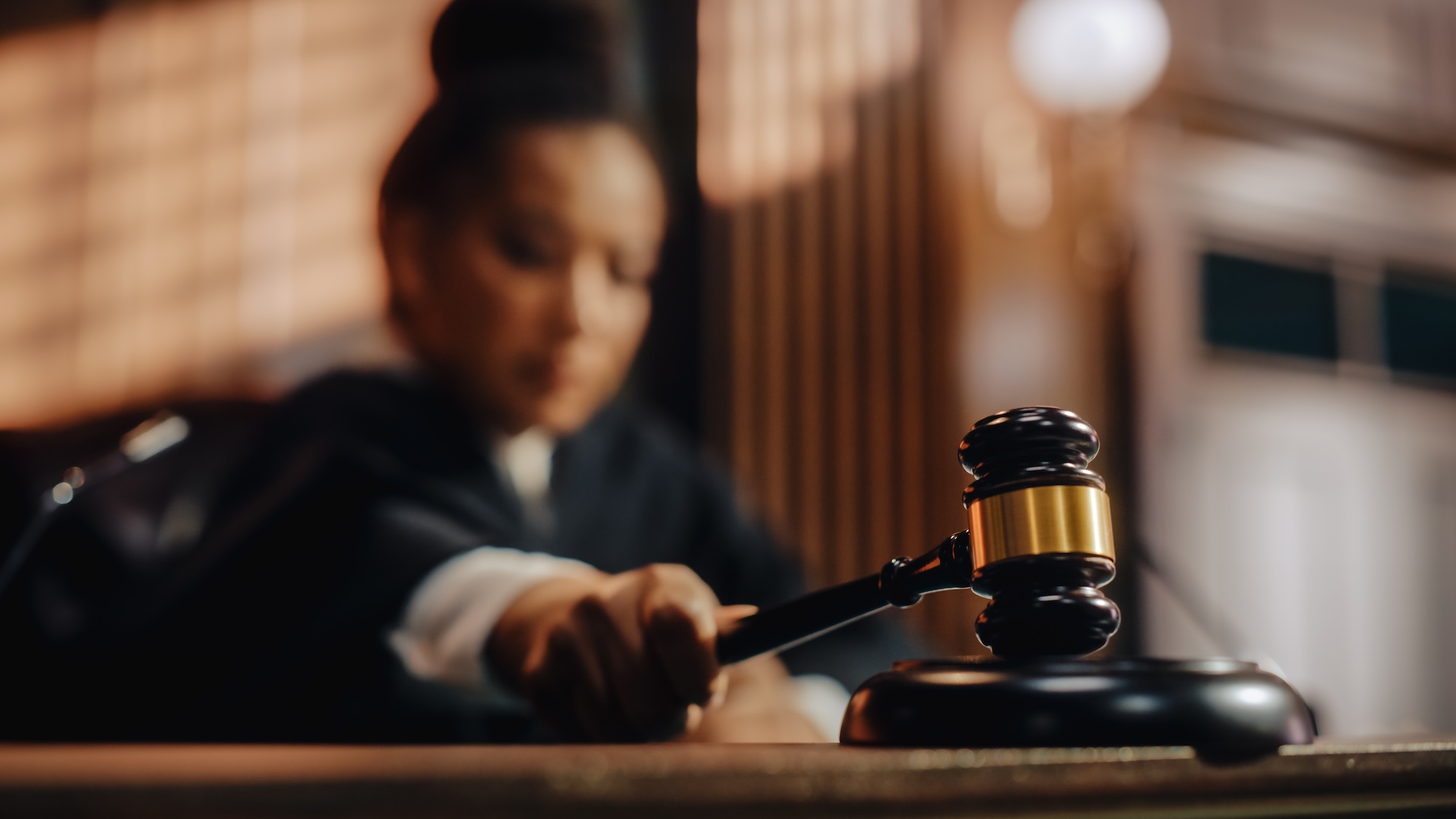 A judge in a courtroom focuses on striking a wooden gavel against a sound block, with the judges face blurred in the background.