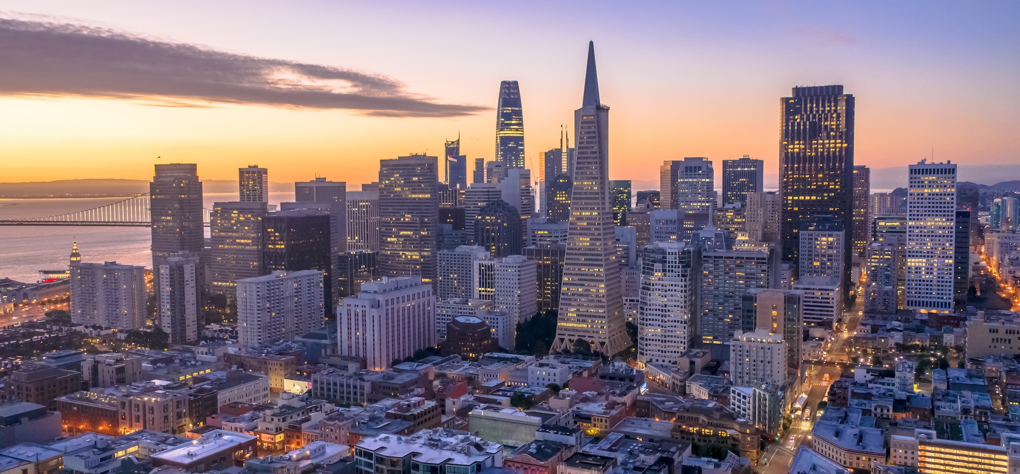 Aerial view of downtown San Francisco at sunset, featuring the Transamerica Pyramid, Salesforce Tower, and other skyscrapers, with the Bay Bridge and calm bay waters in the background.