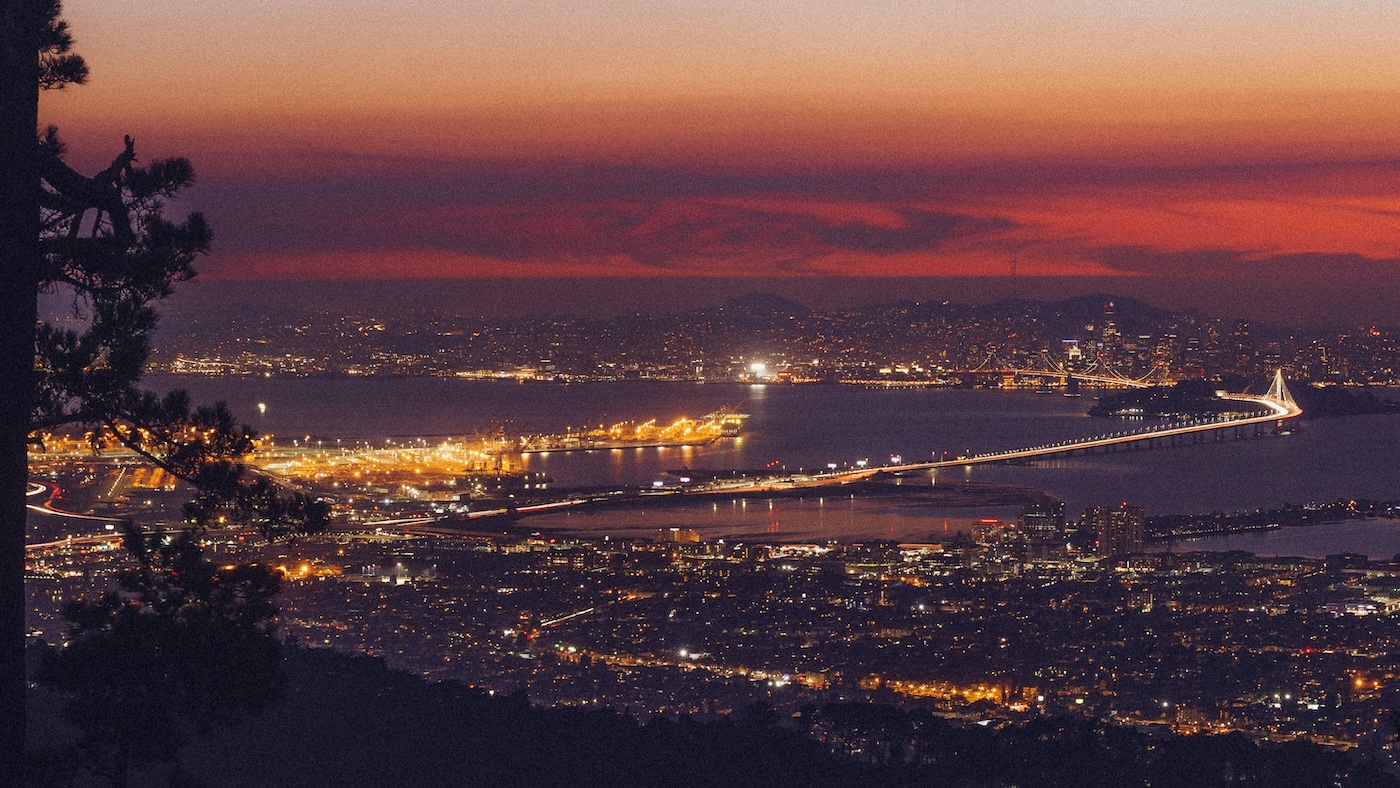 A wide view of a city at sunset, showing a brightly lit bridge stretching over water with city lights, mountains in the background, and a dramatic orange and purple sky.
