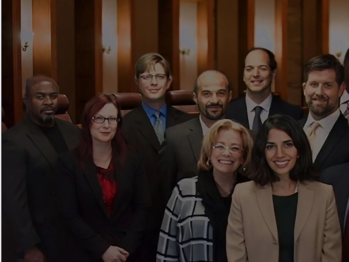 A group of nine professionally dressed adults, both men and women, stand together indoors, smiling at the camera in what appears to be a formal or office setting with wood-paneled walls.