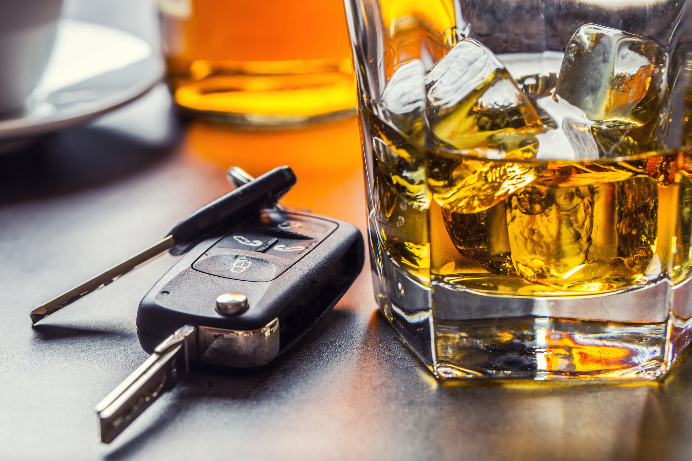 A close-up of a glass of whiskey with ice next to a set of car keys on a table, suggesting the dangers of drinking and driving.