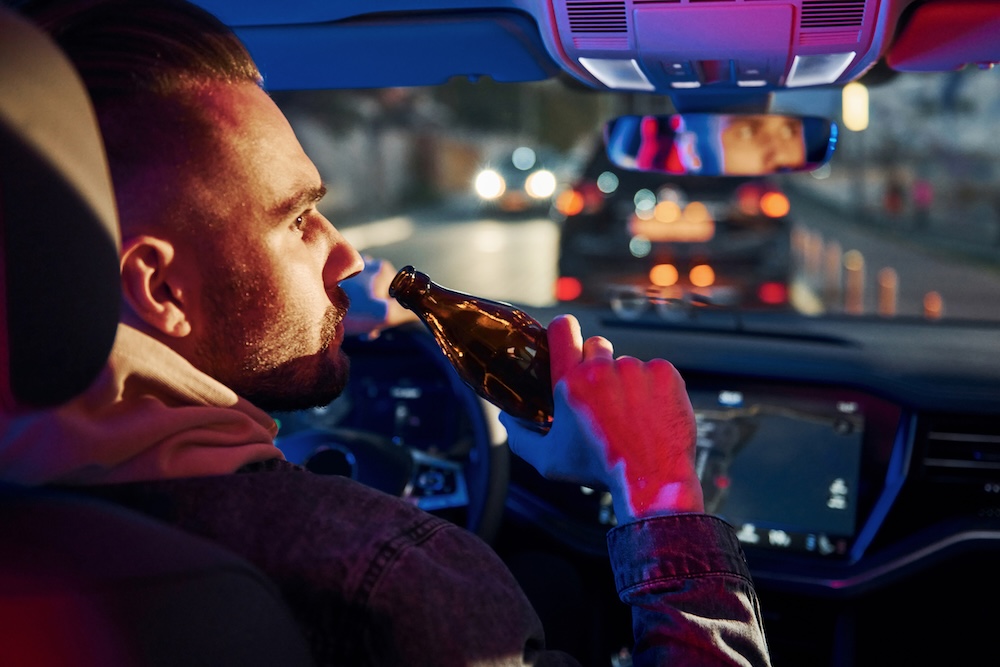 A man sitting in the driver’s seat of a car drinks from a brown bottle, while looking at the road ahead during nighttime traffic.