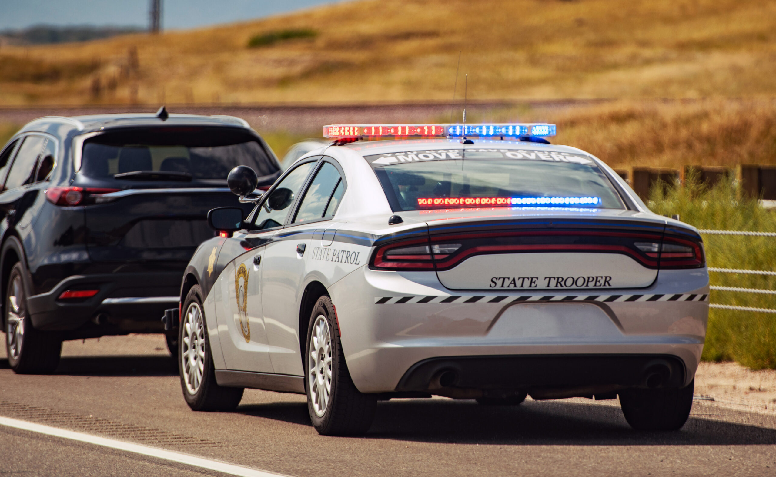 A police state trooper car with flashing lights is parked behind a black SUV on a rural road, likely conducting a traffic stop. Dry grassy fields and a fence are visible in the background.