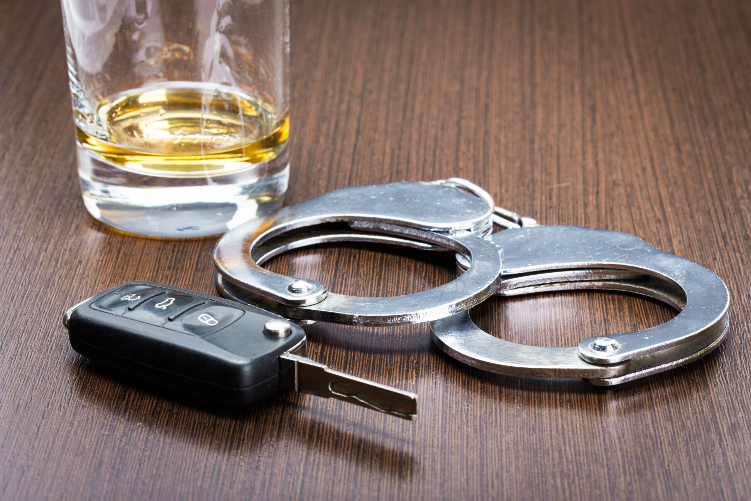 A glass of whiskey, a pair of metal handcuffs, and a car key fob are placed on a wooden table, symbolizing the dangers and consequences of drinking and driving.