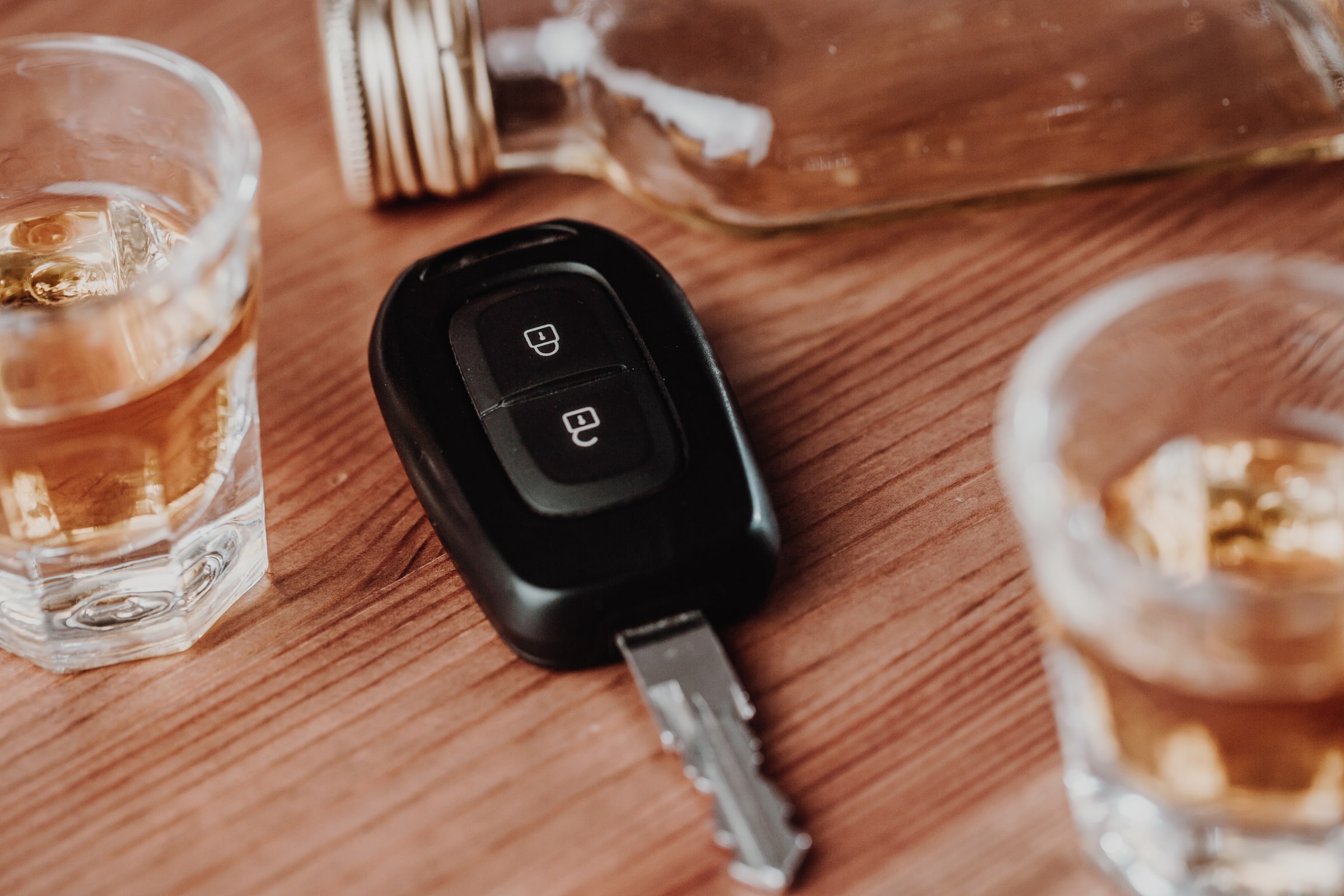 A car key is placed on a wooden table surrounded by two filled shot glasses and a tipped-over bottle of liquor, highlighting the dangers of drinking and driving.