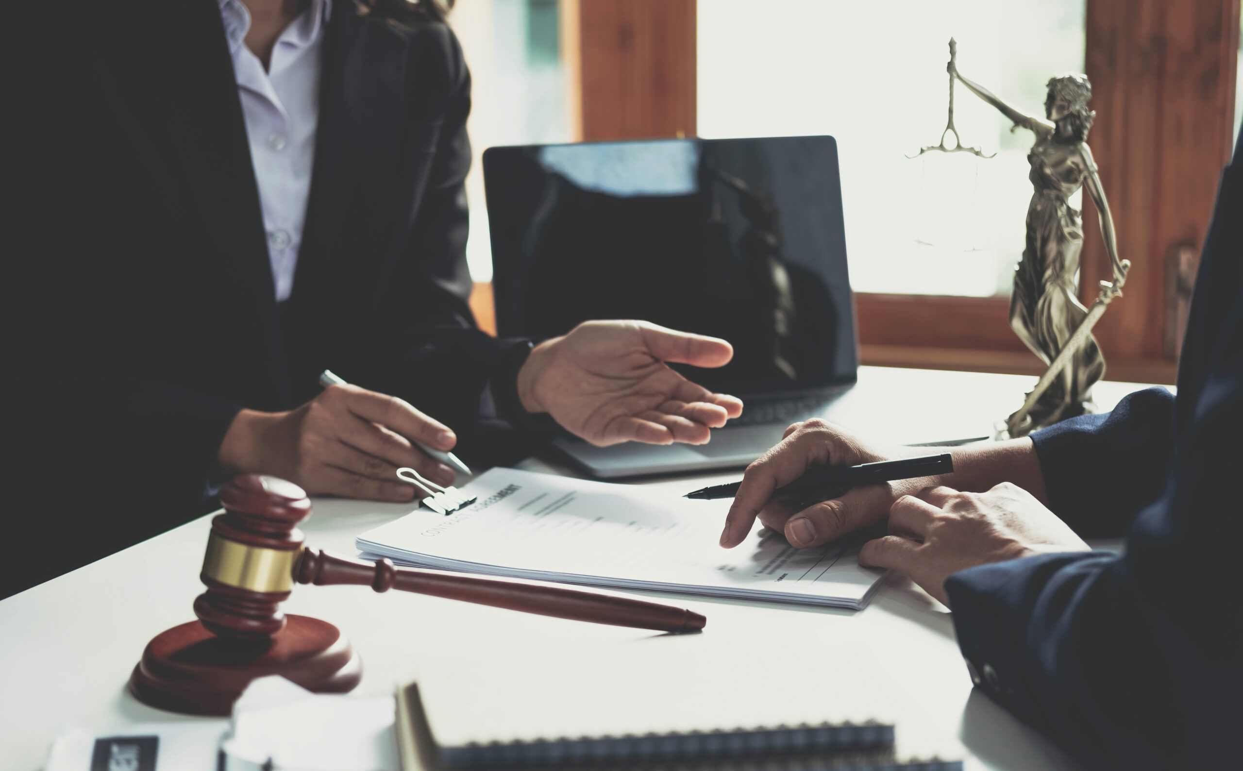 Two people in business suits sit at a desk with legal documents, a gavel, and a scales of justice statue, discussing paperwork in a law office setting.