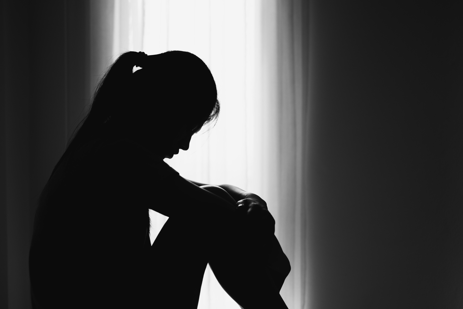 Silhouette of a person sitting with knees drawn to their chest, head bowed, in front of a bright window, conveying a sense of sadness or contemplation.