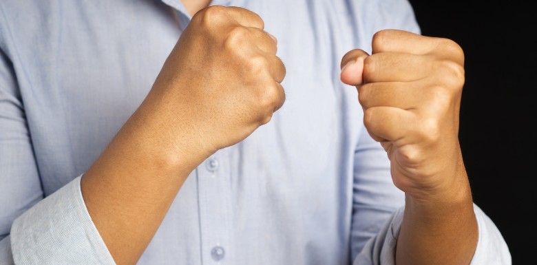 A person in a light blue shirt holds up both hands in clenched fists, positioned as if ready to fight or defend themselves, against a dark background.