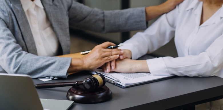 A person in a suit comforts another person by holding their hand at a desk with legal documents, a laptop, and a judge’s gavel, suggesting a supportive legal consultation.