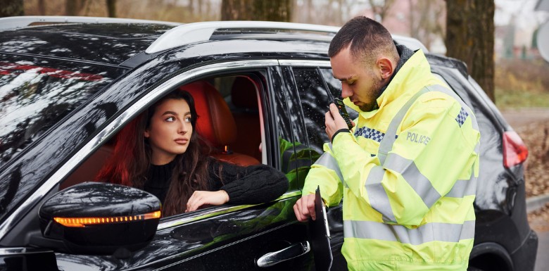 A police officer in a yellow jacket speaks into a radio while standing beside a black car. A woman sits in the drivers seat, looking at the officer through the open window. Trees are visible in the background.