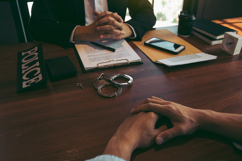 Two people sit across a wooden desk; one has hands clasped and paperwork, with handcuffs, a police badge, and a phone on the table, while the other’s hands are visible in the foreground.