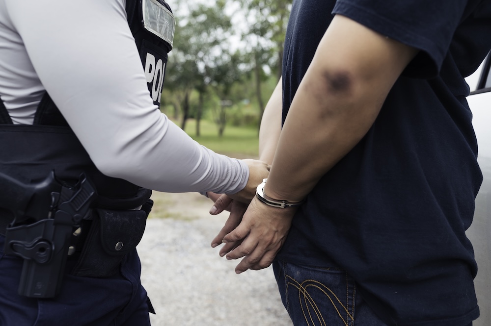 A police officer places handcuffs on a persons wrists behind their back outdoors. The officer is in uniform, and the detained person stands next to a vehicle. Trees and grass are visible in the background.