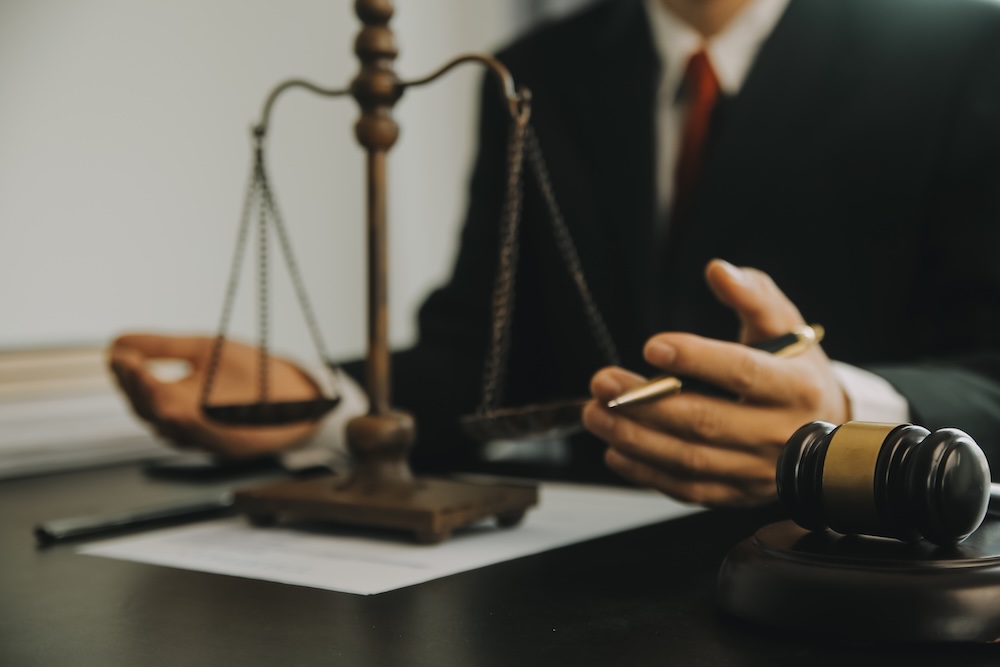 A person in a suit sits at a desk with a balance scale, documents, and a judge’s gavel, suggesting a legal or judicial setting.